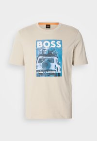 Tricou din bumbac bej, cu un grafic al unui vehicul cu bagaje deasupra și textul "BOSS PAN-AMERICAN" sub imagine.