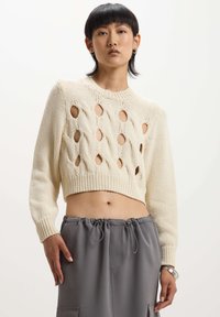 Maglione all'uncinetto di colore crema con motivo a treccia aperta, design cropped e maniche lunghe. Abbinato a pantaloni grigi con cordino.
