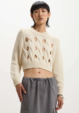 Maglione all'uncinetto di colore crema con motivo a treccia aperta, design cropped e maniche lunghe. Abbinato a pantaloni grigi con cordino.