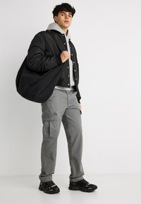 Νεαρός άνδρας με μαύρο μπουφάν, γκρι φούτερ με κουκούλα, παντελόνι cargo και μαύρα αθλητικά παπούτσια, κρατώντας μια μεγάλη μαύρη τσάντα στον ώμο, μπροστά σε λευκό φόντο.