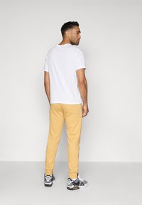 Muž stojaci a pozerajúci sa späť, oblečený v bielom tričku, béžových teplákových nohaviciach, bielych ponožkách a čierno-bielych teniskách na jednoduchom pozadí.