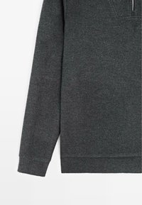 Tmavě šedý pulovr s dlouhými rukávy, vyrobený z měkké, texturované látky. Má půlzip a mírně zaoblený spodní okraj.