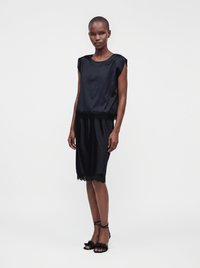 Femme debout portant une robe en satin noire sans manches avec une bordure en dentelle et des talons noirs à brides sur un fond blanc uni.