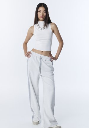 Jonge vrouw staat met handen in de zij, draagt een wit mouwloos cropped topje, wijde sweatpants met zijstrepen, robuuste sneakers en een ketting met een hartje.