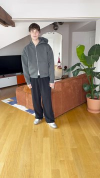 Grauer Kapuzenpullover mit Reißverschluss, schwarze weite Hose, silberne Schuhe; stehend in einem Raum mit Holzboden und einer braunen Couch neben einer grünen Pflanze.