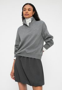 Szürke pulóver, magas gallérral és gombdíszítéssel, sötétszürke, redős szoknyával párosítva, sima textúrával.