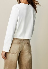 Frau mit langen Haaren, die ein weißes Longsleeve-Oberteil und hochtaillierte beige Jeans trägt, steht vor einem schlichten Hintergrund.
