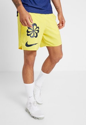 Mężczyzna ubrany w żółte spodenki Nike z czarnymi kwiatowymi wzorami i logo Swoosh, białe skarpetki Nike oraz białe sneakersy, stojący na białym tle.