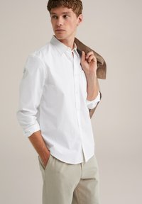 Witte button-up shirt met opgerolde mouwen, gecombineerd met lichtbeige broeken. Het shirt heeft een klassieke kraag en een borstzak.