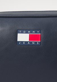 Tamnoplava kožna torba s pravokutnim logotipom u crvenoj, bijeloj i plavoj boji; uključuje urezani natpis "TOMMY JEANS." Tekstura površine.