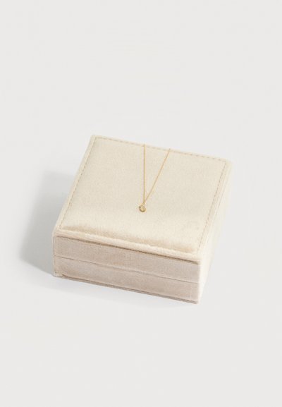 Κολιέ με χρυσή αλυσίδα και μικρό στρογγυλό μενταγιόν τοποθετημένο σε κλειστό μπεζ βελούδινο κουτί κοσμημάτων πάνω σε ανοιχτόχρωμο φόντο.