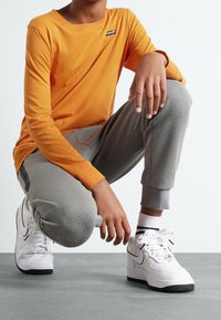 Orange långärmad skjorta med en liten logotyp, grå texturerade joggers och vita sneakers med svarta accenter, med en lågt skuren design.