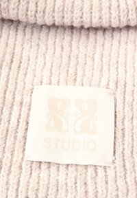 Tejido de punto beige claro con textura acanalada. Una etiqueta blanca presenta un logo sutil y la palabra "STUDIO" en letras en relieve.