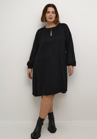 Robe noire à manches longues avec un col rond et un détail en forme de trou de clé. Tissu lisse et fluide avec des accents froncés au niveau du col et des poignets.