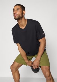 Svart bomull T-skjorte med korte ermer, med en logo på venstre erme. Kombinert med olivengrønne shorts. Atletisk bygget person som holder en kettlebell.