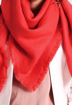 Écharpe texturée rouge vif avec bords à franges enroulée autour du cou, portée sur un haut rose clair et une veste blanche.