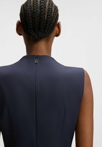 Merengek sleeveless dress dengan leher tinggi dan zipper tersembunyi di belakang; kain halus dengan potongan yang pas.