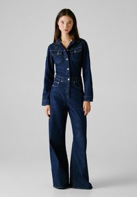 Menton bleu foncé en denim avec des manches longues, col, fermeture à boutons, deux poches poitrine et coupe ample. Texture lisse et taille cintrée.