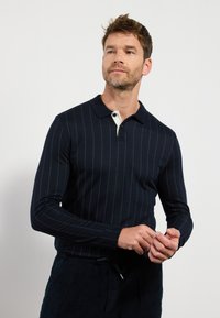 Marineblå langærmet polo med vertikale hvide striber, krave med cremefarvet detalje, der har et tætsiddende design og ribbet tekstur.