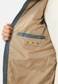 Gepolsterte Jacke mit blauer Außenseite und beiger Innenauskleidung. Verfügt über eine kleine Tasche und Symbole, die wasserabweisend, winddicht und daunenfrei anzeigen.