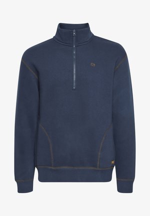 Sötétkék pulóver félszippes gallérral, kontrasztos varrással és kis logóval a mellrészen. Puha anyag, bordás mandzsettákkal és szegélyekkel.