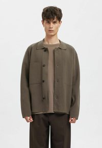 Nadměrná olivově zelená bunda s límcem, zapínáním na knoflíky a dvěma předními kapsami. Nositelná přes béžové tričko a hnědé kalhoty.