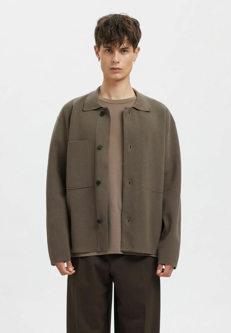 Nadměrná olivově zelená bunda s límcem, zapínáním na knoflíky a dvěma předními kapsami. Nositelná přes béžové tričko a hnědé kalhoty.