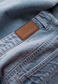 Jeans de mezclilla azul claro con un primer plano del bolsillo trasero y una etiqueta de cuero marrón que dice "ombre" cosida encima del bolsillo.