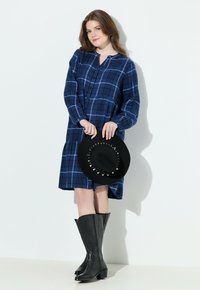 Kvinna i blå rutigt klänning och svarta cowboyboots håller en svart hatt med stardekorationer mot vit bakgrund.
