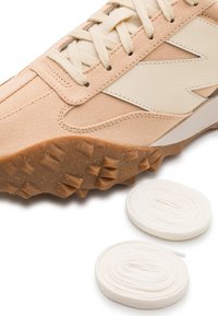 Beige sneaker with textured gum sole and white zigzag detail, accompanied by two coiled white shoelaces on a white background.