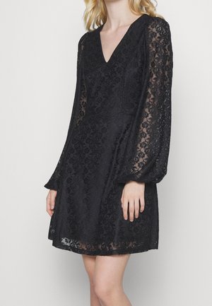 Donna che indossa un vestito nero in pizzo con maniche lunghe trasparenti e scollatura a V, in piedi davanti a uno sfondo chiaro e semplice.