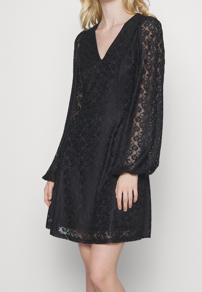 Femme portant une robe en dentelle noire avec de longues manches transparentes et un décolleté en V, debout devant un fond clair uni.