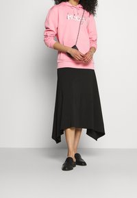 Donna vestita con un hoodie rosa, una borsa a tracolla nera, una gonna midi asimmetrica nera e mocassini neri, in piedi contro uno sfondo neutro.