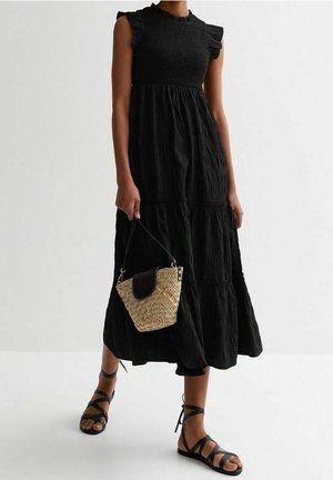 Kobieta ubrana w czarną, bezrękawnikową sukienkę midi z falbanami na ramionach, trzymająca plecioną słomianą torbę i czarne płaskie sandały na paski.