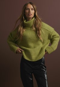 Rohke roheline kaelus-sweater, ribakujundusega, ülelaia lõikega, langevate õlgadega, kombineeritud läikivate mustade pükstega pruunil taustal.