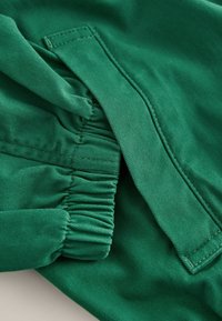 Tissu vert avec un poignet élastique froncé et un rabat rectangulaire fixé par couture, montrant un gros plan détaillé de la couture et de la texture.