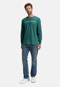 Maglietta a maniche lunghe in teal con logo bianco "TOM TAILOR", abbinata a jeans blu e sneakers bianche. Vestibilità casual con un aspetto confortevole.