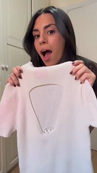 Frau mit dunklen Haaren hält ein weißes, kurzärmeliges, geripptes Shirt mit großem Ausschnitt und silberner Schnalle in einem Raum.