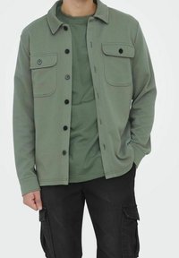 Chaqueta verde oliva con tejido texturizado, cierre de botones al frente, dos bolsillos en el pecho, llevada sobre una camisa verde a juego y pantalones cargo negros.