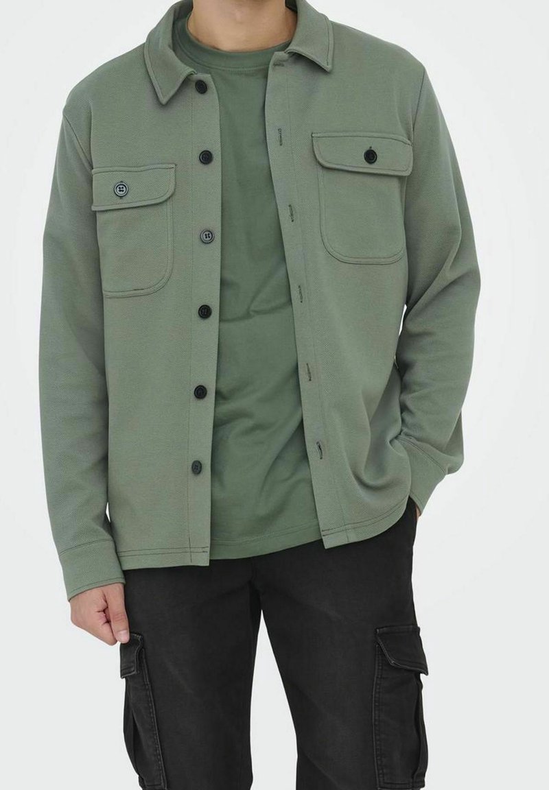 Chaqueta verde oliva con tejido texturizado, cierre de botones al frente, dos bolsillos en el pecho, llevada sobre una camisa verde a juego y pantalones cargo negros.