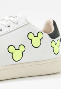 Białe skórzane sneakersy z haftowanymi neonowo-żółtymi sylwetkami Myszki Mickey, czarnym akcentem na pięcie oraz teksturowaną gumową podeszwą.
