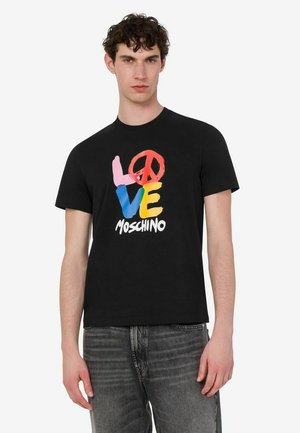 Młody mężczyzna w czarnej koszulce z kolorowym "LOVE" i grafiką znaku pokoju oraz tekstem "Moschino", w połączeniu z wyblakłymi czarnymi dżinsami.