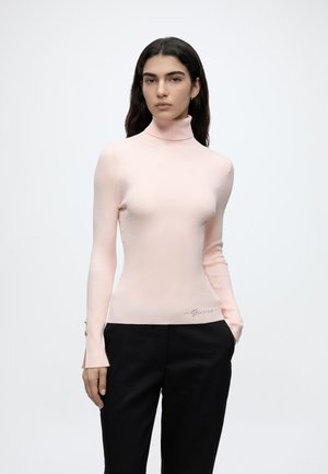 Femme aux longs cheveux foncés porte un pull à col roulé rose pâle et un pantalon noir, debout, une main dans la poche, sur un fond uni.