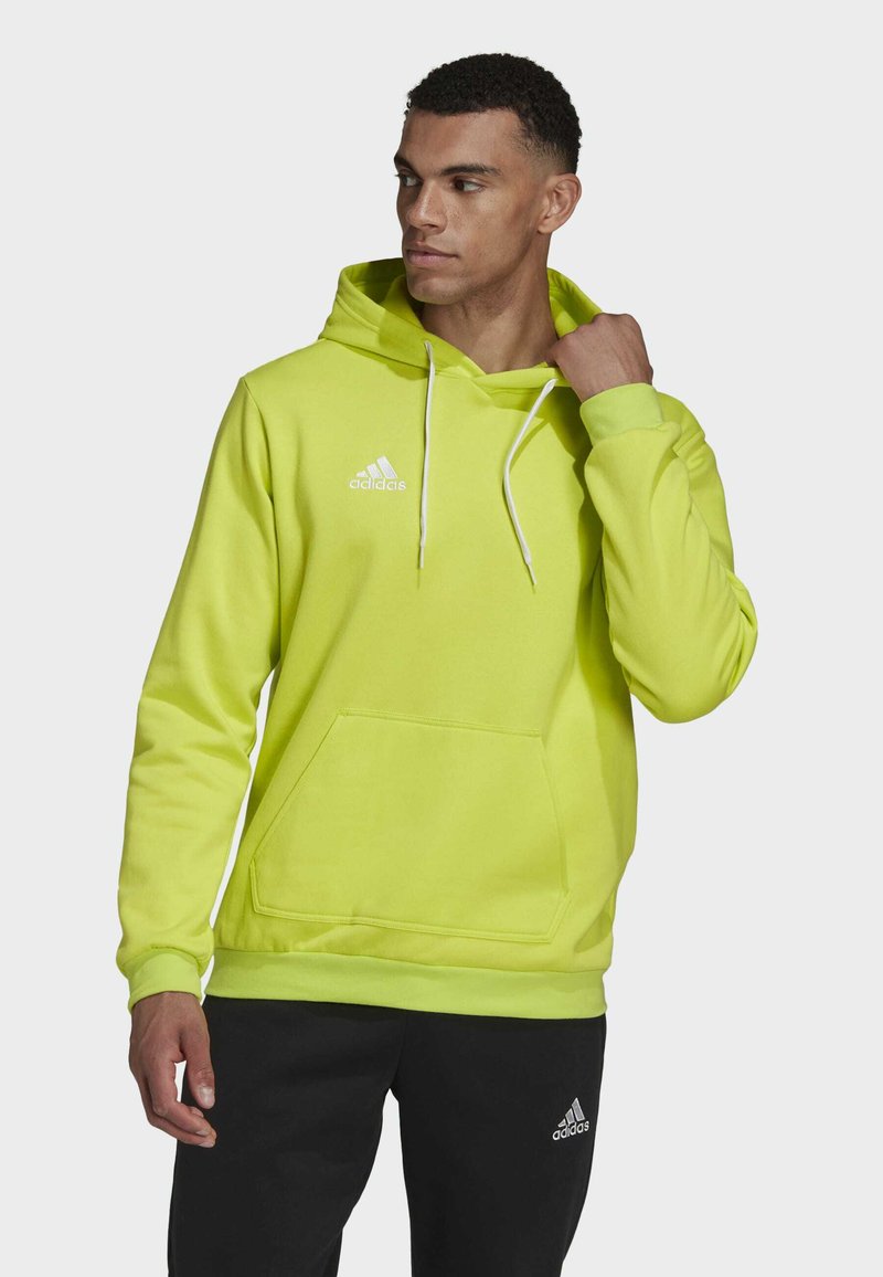 Neon-gelbes Hoodie mit einer vorderen Tasche und weißen Kordeln, ausgestattet mit einem gestickten Logo auf der Brust und gerippten Bündchen. Kombiniert mit schwarzen Hosen.