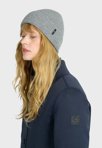 Donna con lunghi capelli biondi che indossa un berretto di maglia grigio e una giacca blu scuro con una toppa a forma di foglia d'acero canadese sulla manica.