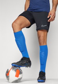 Pantaloni sportivi neri con un design aderente, abbinati a calze al ginocchio di colore blu brillante. Scarpini da calcio neri su una palla con motivo bianco e nero.