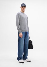 Felpa grigia, jeans blu a gamba larga, sneakers bianche e una borsa nera con cerniere. Uomo con un cappellino scuro su uno sfondo neutro.