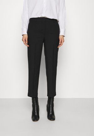 Personne portant un pantalon noir fuselé, des bottines noires et une chemise blanche à manches longues boutonnée sur un fond blanc uni.