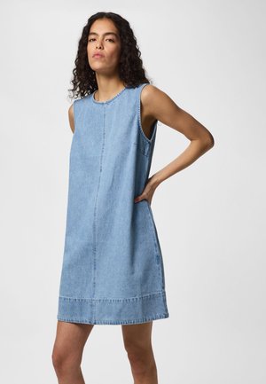 Γυναίκα με σγουρά μαλλιά που φοράει αμάνικο ανοιχτό μπλε φόρεμα τζιν, στέκεται με το ένα χέρι στη μέση σε μια μονήχρωμη φόντο.