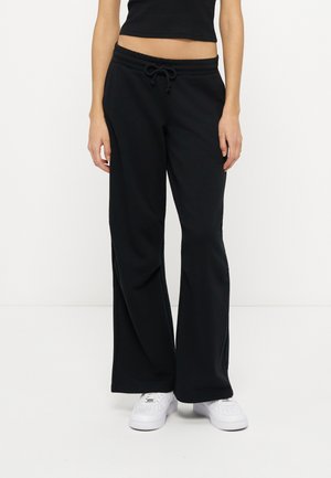 Femme portant un pantalon noir à jambes larges avec taille à cordon et des baskets blanches, debout devant un fond blanc uni.